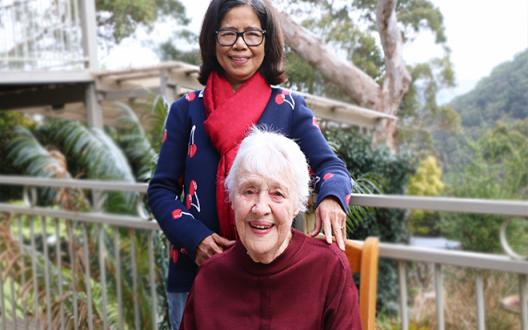 rebecca in a purple knit jumper and yvonne in a red sweater smiling for a photo on a balcony by the bush